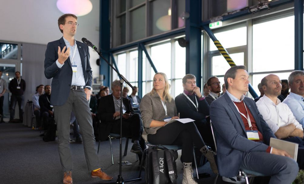 Mann stehend auf einem Gang spricht in einem Konferenzraum in ein Mikrofon, neben ihm sitzendes Publikum